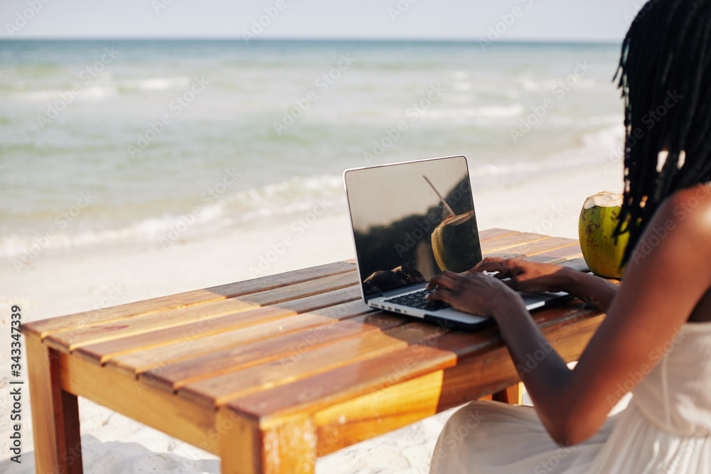 Female freelancer or blogger sitting at table on the beautiful beach and working on laptop