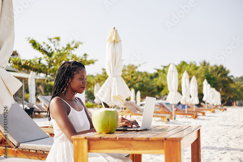 Pretty young woman sitting at table on sandy beach, enjoying fresh coconut cocktail and working on laptop photo