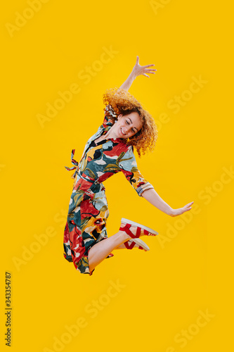 Happy exhilarated woman in a colorful dress jumping in the air over yellow. Arms bent, arms outstretched. taking artistic form © zzzdim
