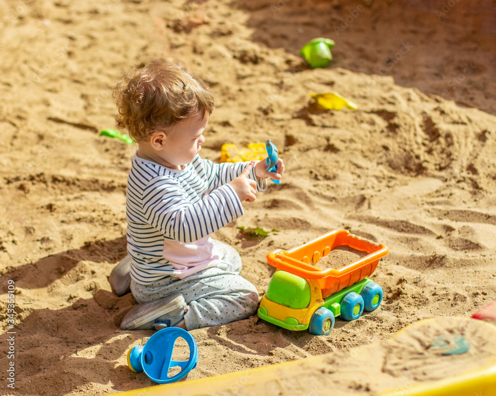 Little baby girl Caucasian boy plays in the sandbox with a toy car. A child in a striped t-shirt pours sand into a truck with a spatula Little baby girl Caucasian boy plays in the sandbox with a toy car. A child in a striped t-shirt pours sand into a truck with a spatula