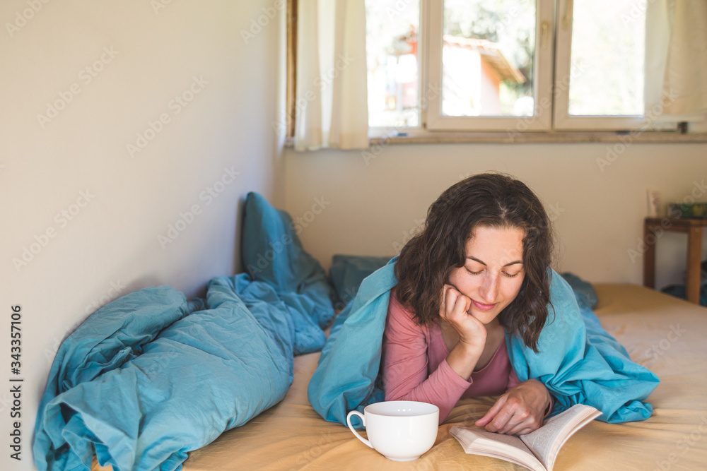 Smiling girl reads a book and drinks coffee while lying in bed. Smiling girl reads a book and drinks coffee while lying in bed.
