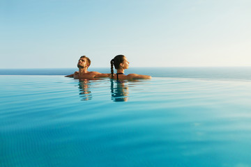 Couple In Love At Luxury Resort On Romantic Summer Vacation. People Relaxing Together In Edge Swimming Pool Water, Enjoying Beautiful Sea View. Happy Lovers On Honeymoon Travel. Relationship, Romance