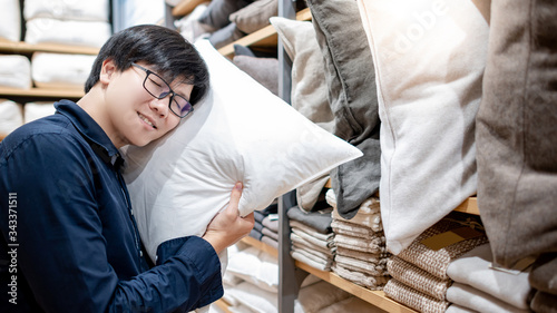 Young Asian man customer hugging comfortable pillow choosing for his bedroom in furniture store. Shopping house decoration for interior room design. Home improvement concept