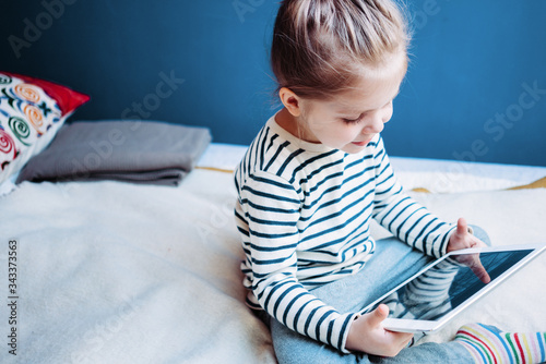 Cute kid watching cartoons at home using wireless tablet computer. Girl toddler growing with gadget in cozy place