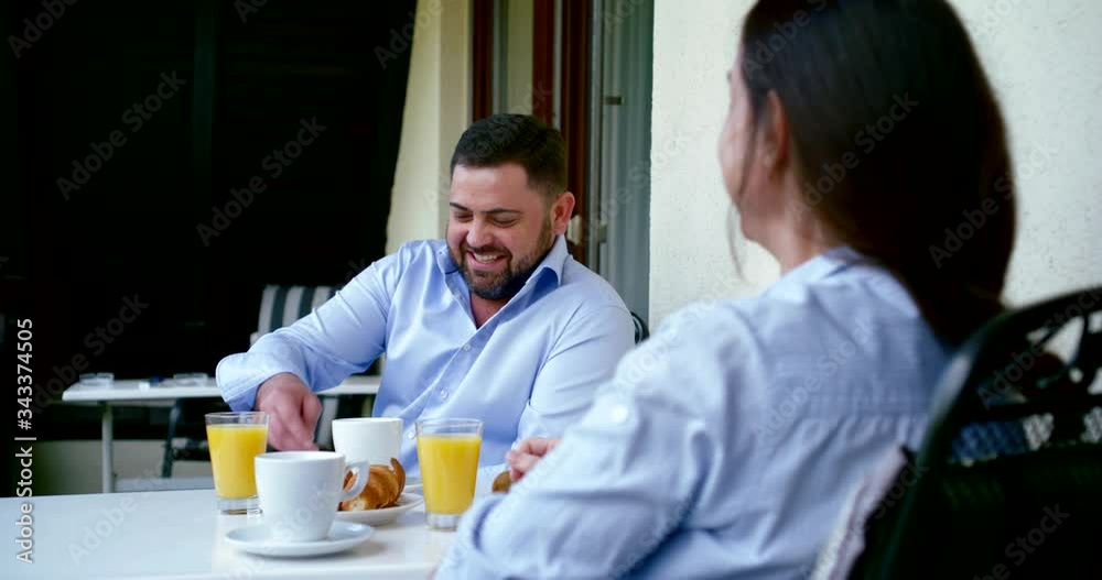 a couple sits on a balcony at a table with coffee, juice and fruit. they are talking. in the foreground a woman straightening her hair. in the background a man touches a saucer and laughs a couple sits on a balcony at a table with coffee, juice and fruit. they are talking. in the foreground a woman straightening her hair. in the background a man touches a saucer and laughs