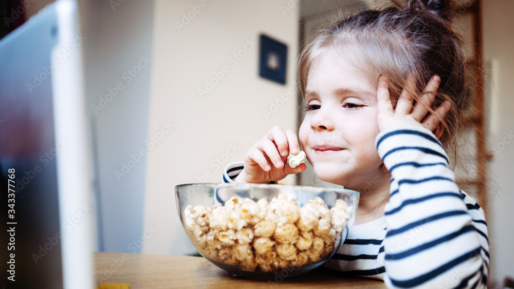 Smiling kid watching cartoons at home using wireless tablet computer and eating pop-corn. Girl toddler growing with gadget in cozy place Smiling kid watching cartoons at home using wireless tablet computer and eating pop-corn. Girl toddler growing with gadget in cozy place