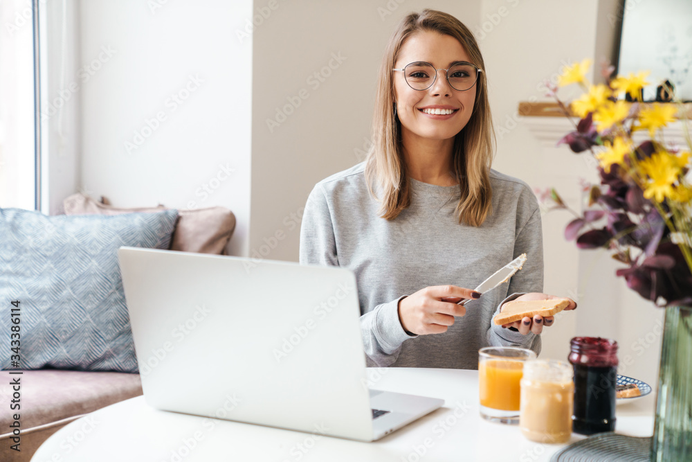 Photo of cheerful woman in eyeglasses eating toasts while using laptop Photo of cheerful woman in eyeglasses eating toasts while using laptop