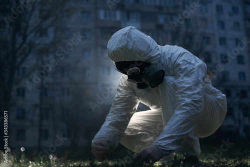 Close-up snapshot of a crouching researcher working outside with fresh samples using laboratory tools and liquids, concept of saving our planet. Ecologist wearing white uniform, gloves and gas mask