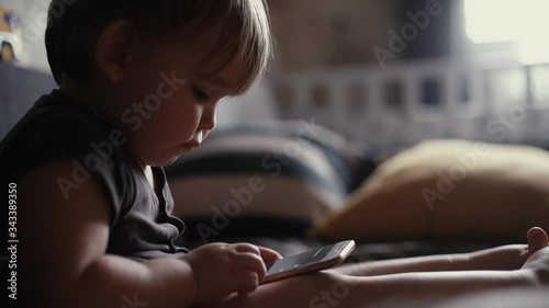 Adorable chubby child boy touching screen display smartphone, playing with phone while sitting on soft couch in living room. Concept of modern entertainment young children. Shooting in slow motion. photo