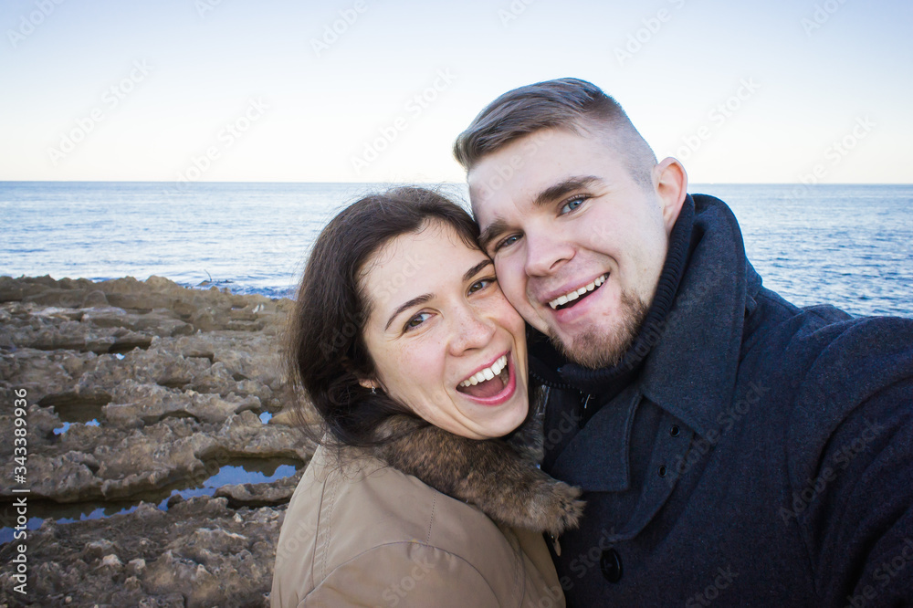 Portrait of young beautiful couple taking selfie photo in a smartphone with sea and dark cloudy sky on the background. Cold season and travel concept.