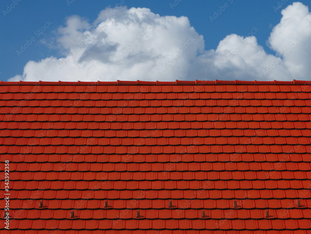 Red clay tile sloped roof and ridge. bright blue sky and fluffy white clouds. modern residential construction materials concept. abstract low angle view. metal snow and ice breakers along the bottom Red clay tile sloped roof and ridge. bright blue sky and fluffy white clouds. modern residential construction materials concept. abstract low angle view. metal snow and ice breakers along the bottom