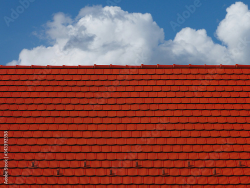 Red clay tile sloped roof and ridge. bright blue sky and fluffy white clouds. modern residential construction materials concept. abstract low angle view. metal snow and ice breakers along the bottom 