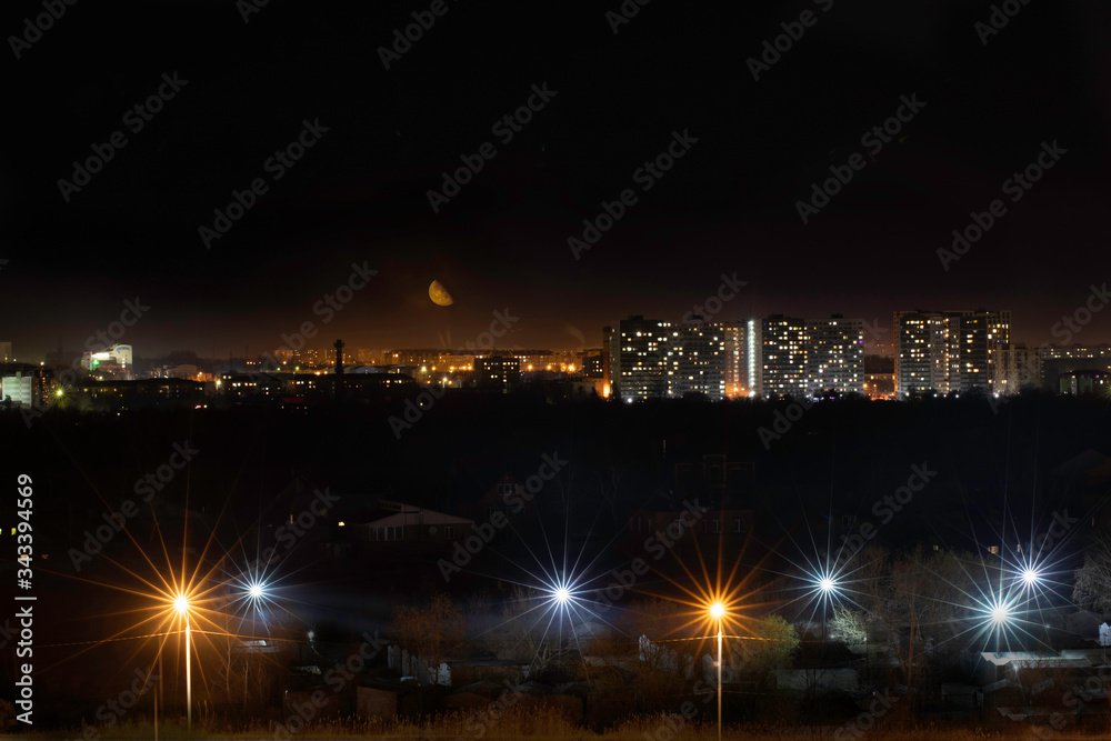 Panoramic view of night city. Lights of multistoried buildings on background, bright street lamps in the forefront. Urbanistic concept, city lights, mysterious places, moonlighting, hidden in darkness