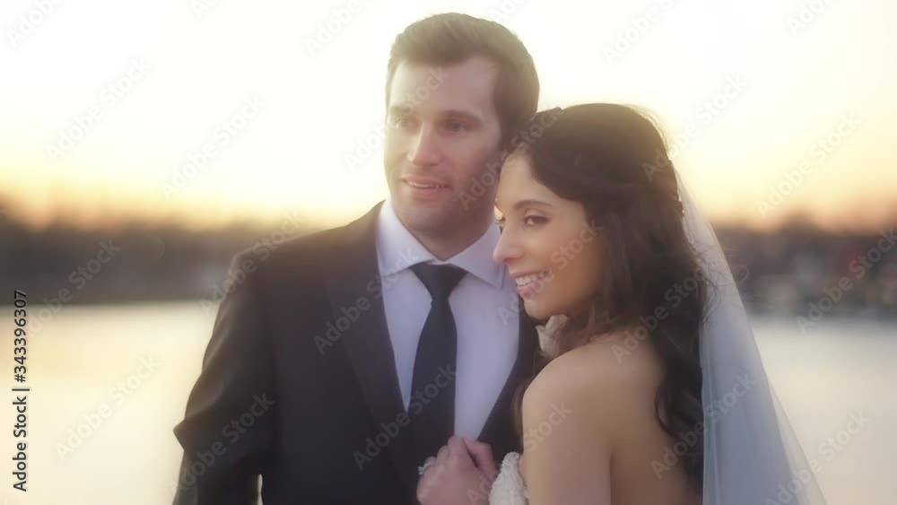 A bride and groom stand close and smile together as the haze of the sunset illuminates them and the river backdrop A bride and groom stand close and smile together as the haze of the sunset illuminates them and the river backdrop