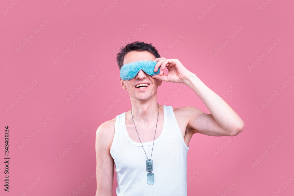 Cheerful positive young skinny man with sleeping mask on face touch it with hand. Posing on camera alone in studio. Isolated over pink background.