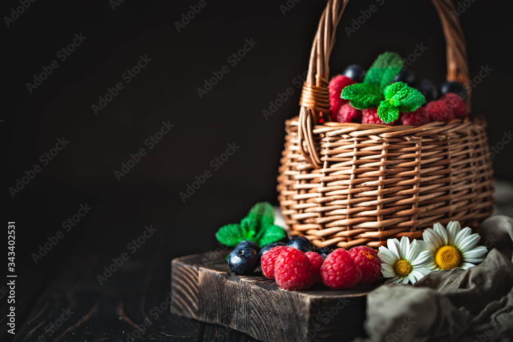 Raspberries and blueberries in a basket with chamomile and leaves on a dark background. Summer and healthy food concept. Background with copy space. Selective focus. Raspberries and blueberries in a basket with chamomile and leaves on a dark background. Summer and healthy food concept. Background with copy space. Selective focus.
