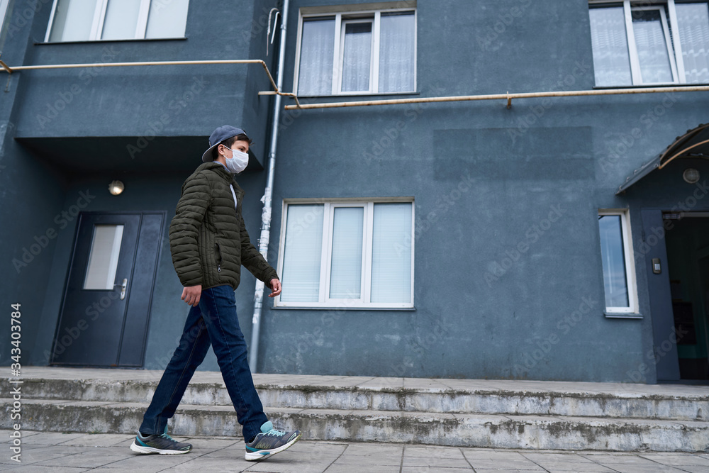 teen boy walks near wall of high-rise buildings with apartments, a residential area, a medical mask on his face protects against viruses and dust teen boy walks near wall of high-rise buildings with apartments, a residential area, a medical mask on his face protects against viruses and dust