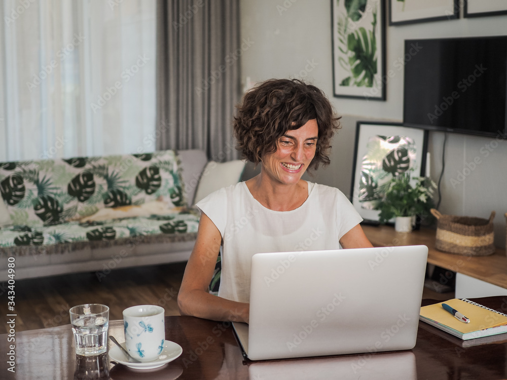 happy smiling remote online working woman with laptop, notebook and glass in casual outfit sitting on a work desk in her living room in her home office happy smiling remote online working woman with laptop, notebook and glass in casual outfit sitting on a work desk in her living room in her home office