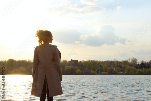 Blurry image of teenage girl wearing a beige coat and blue jeans standing near the water, overexposed shot.  People, travel, nature concept.