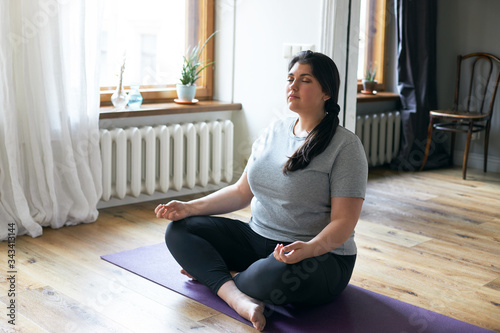 Portrait of plus size young Caucasian woman in sportswear sitting on floor using yoga mat while practicing meditation, keeping legs crossed and eyes closed, having calm peaceful facial expression