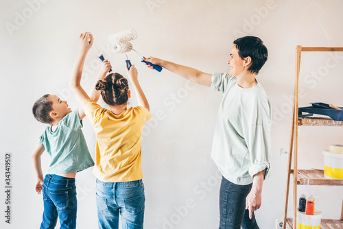 Happy mother, douther and son painting wall with roller. Mom teachs her children painting with roller at home. photo