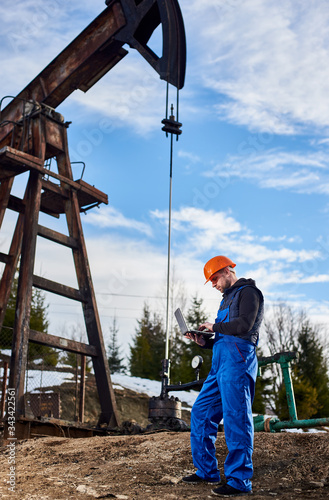 Full length view of male worker using laptop, controlling work of pump jack. Wearing blue uniform and orange helmet. Concept of petroleum industry and oil extraction.