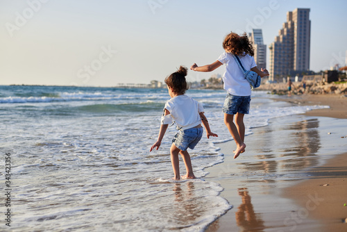 Two small girls sisters, playing jumping on the beach at sunset. Beautiful seaside landscape picture. Family vacation at tropical resort in summer.