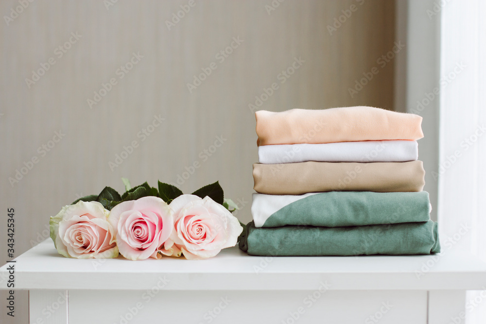 a stack of neatly folded clothes in various muted colors on a white dresser near a light window with a bouquet of pink roses a stack of neatly folded clothes in various muted colors on a white dresser near a light window with a bouquet of pink roses