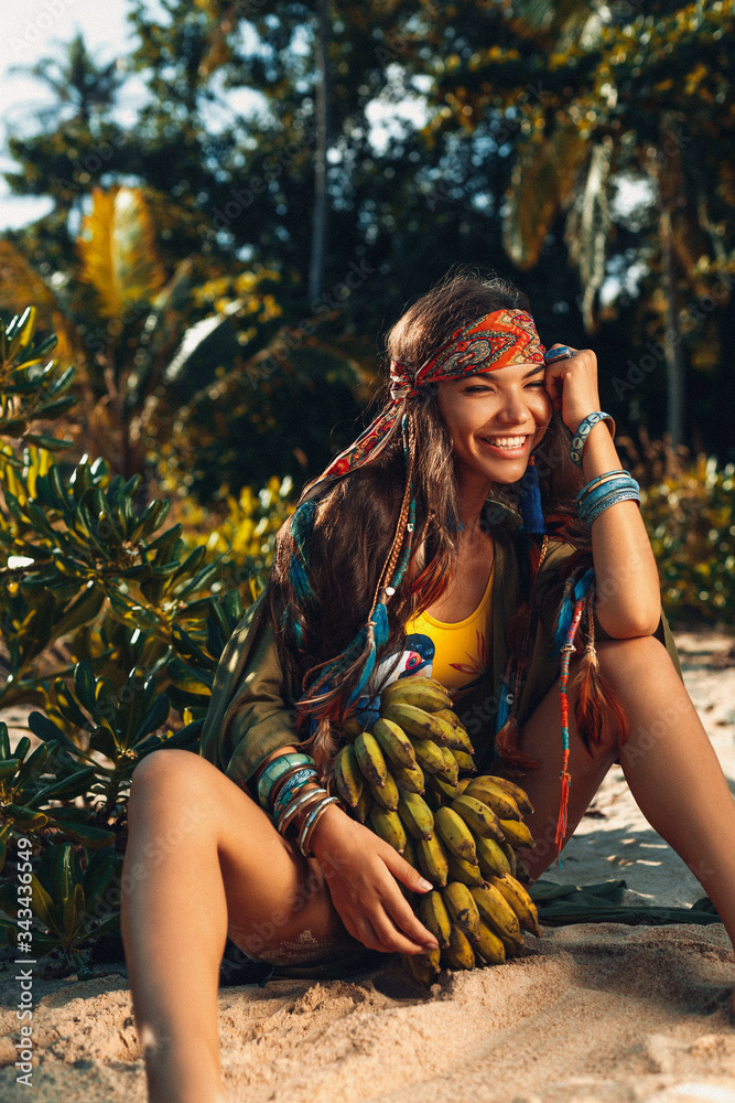 Φωτογραφία cheerful young stylish woman holding a bunch of banans on the feild at sunset