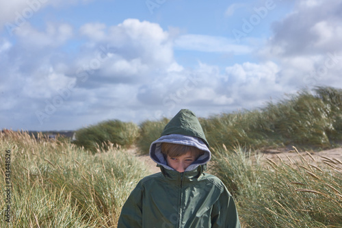 Boy in sand dunes in Denmark, hiding his face in green jacket to protect himself from the cold north wind while looking around, in the background a northern area