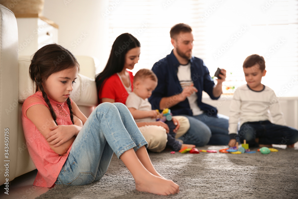 Unhappy little girl feeling jealous while parents spending time with other children at home Unhappy little girl feeling jealous while parents spending time with other children at home
