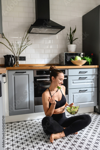 Young woman in sportswear is eating salad sitting on the floor in the kitchen. Healthy lifestyle concept photo