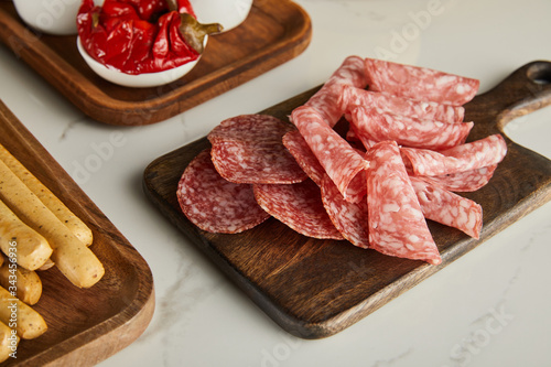 High angle view of salami slices, breadsticks and bowl with marinated chili peppers on boards on white