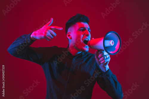 Shouting with speaker. Caucasian young man's portrait isolated on red studio background in neon light. Beautiful male model. Concept of human emotions, facial expression, sales, ad, youth culture.