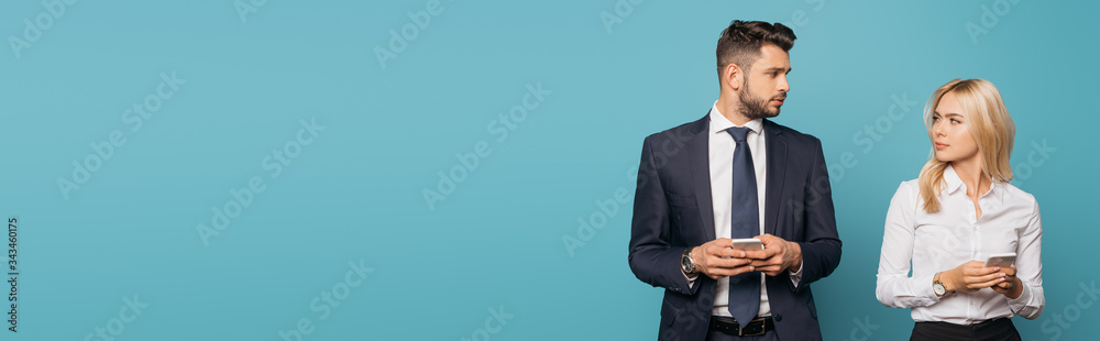 panoramic orientation of young businesswoman and businessman looking at each other while holding smartphones on blue background panoramic orientation of young businesswoman and businessman looking at each other while holding smartphones on blue background