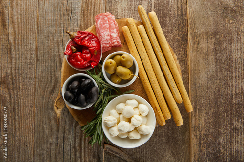 Top view of breadsticks, salami slices and rosemary near bowls with antipasto ingredients on round board on wooden background