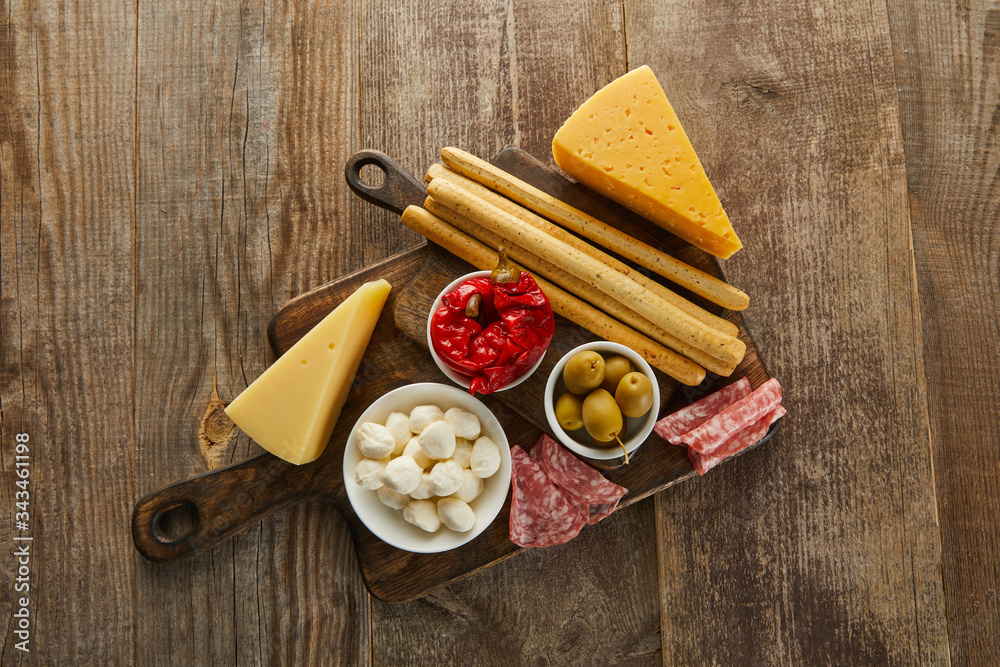 Top view of bowls with antipasto ingredients near breadsticks, salami slices and cheese on boards on wooden background