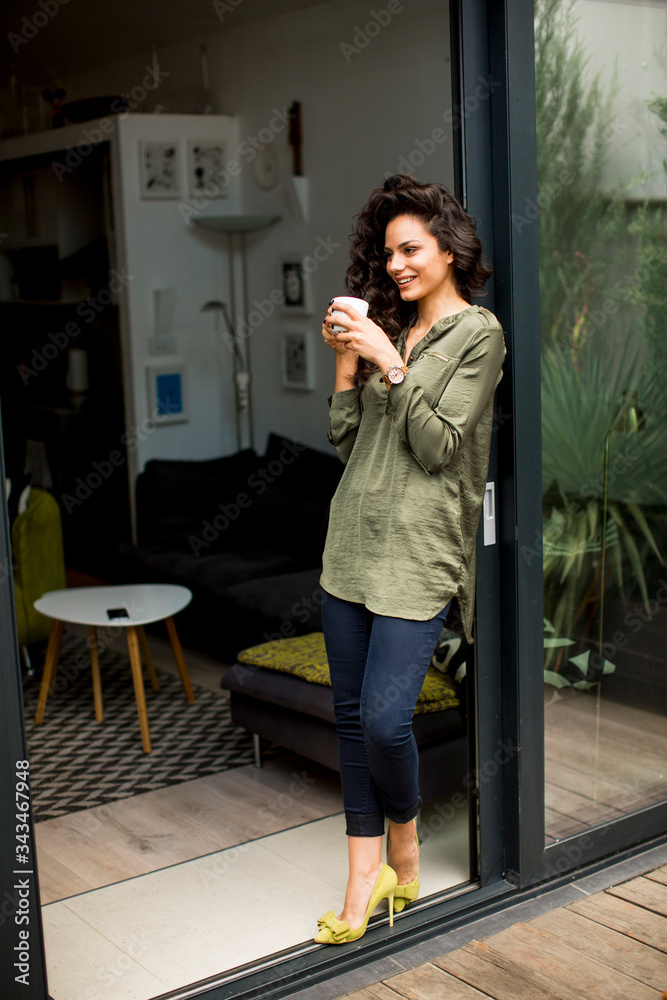 Fototapeta premium Young curly hair woman looking out of window at home backyard and drinking cup of coffee