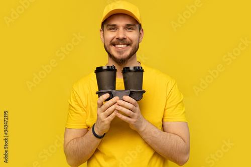 Portrait of cheerful delivery man in yellow uniform smiling and holding takeaway coffee cups isolated over yellow