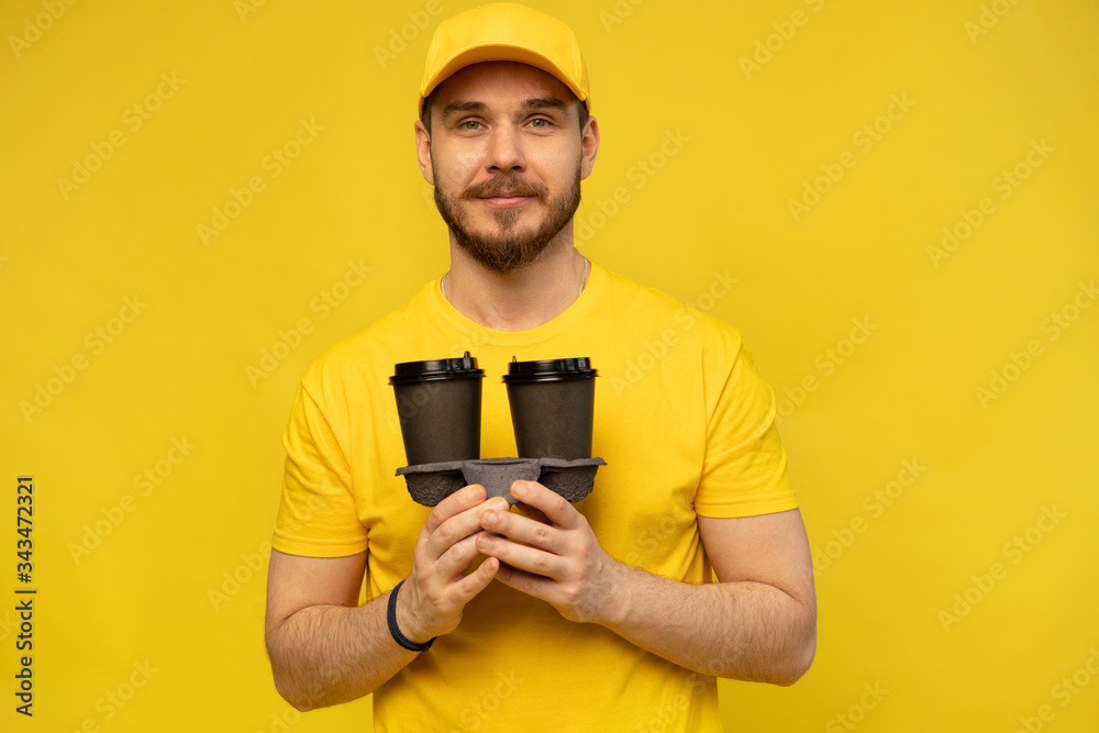 Portrait of cheerful delivery man in yellow uniform smiling and holding takeaway coffee cups isolated over yellow Portrait of cheerful delivery man in yellow uniform smiling and holding takeaway coffee cups isolated over yellow