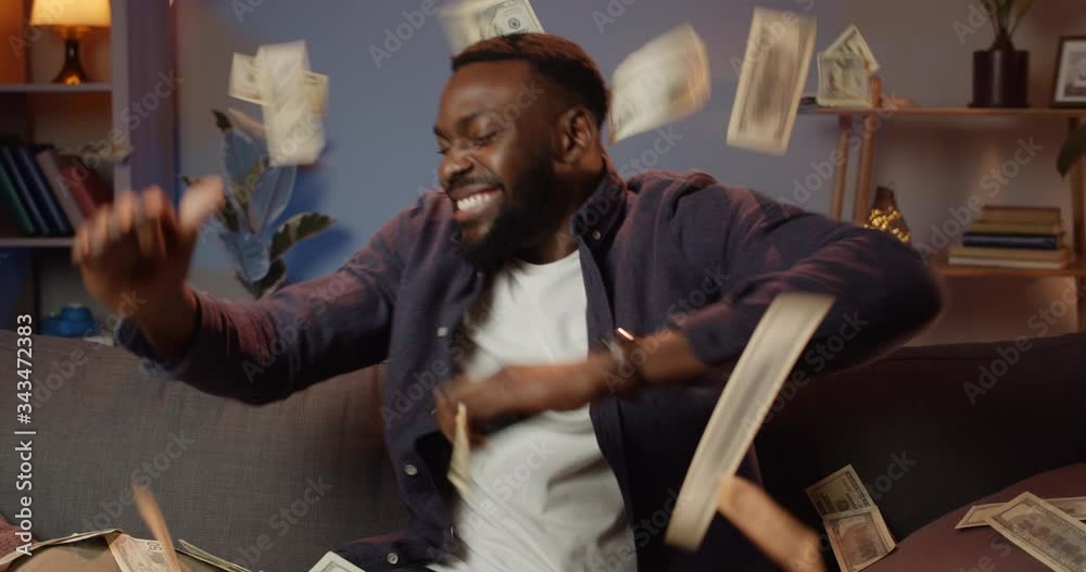 Happy afro american man holding smartphone and making successful deal while working from home. Young guy winning big money and rejoicing. Money falling from above