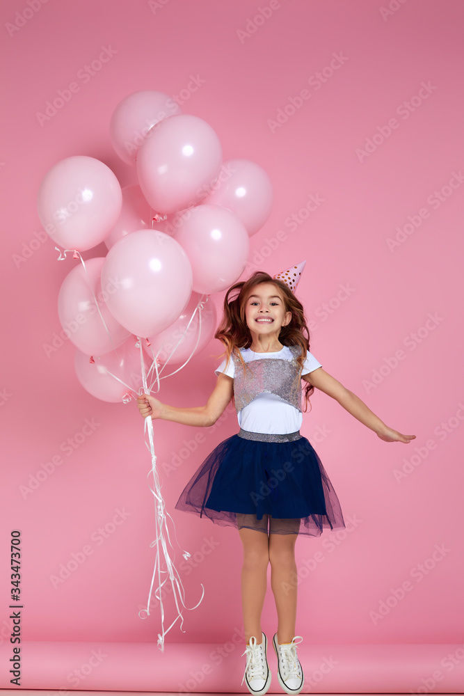 Beautiful happy little child girl in dress and birthday hat jumping with pastel pink air balloons isolated on pink background. birthday party. Beautiful happy little child girl in dress and birthday hat jumping with pastel pink air balloons isolated on pink background. birthday party.