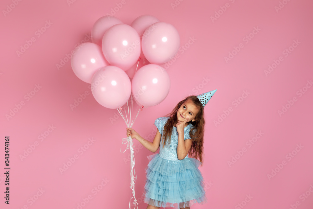 adorable smiling little child girl in blue dress and birthday hat with pastel pink air balloons thinking about something on pink background. birthday party.