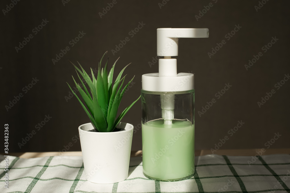 transparent dispenser with green liquid soap stands on a shelf on a white towel with an artificial flower at home in the sun transparent dispenser with green liquid soap stands on a shelf on a white towel with an artificial flower at home in the sun