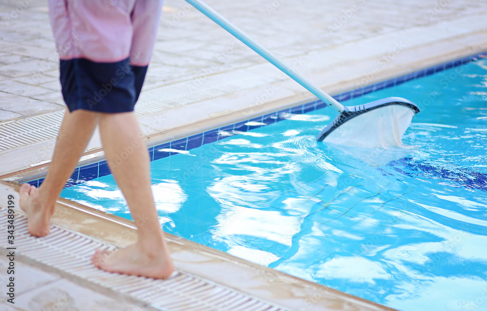 Man cleaning a swimming pool in summer with a brush or net on a blue pole standing barefoot on the tiles at the edge, low angle view of his lower body.