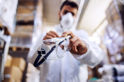 Young man in protective suit putting protective glasses and preparing himself for disinfection from corona virus / covid-19. Warehouse interior. Warehouse is full of food products.