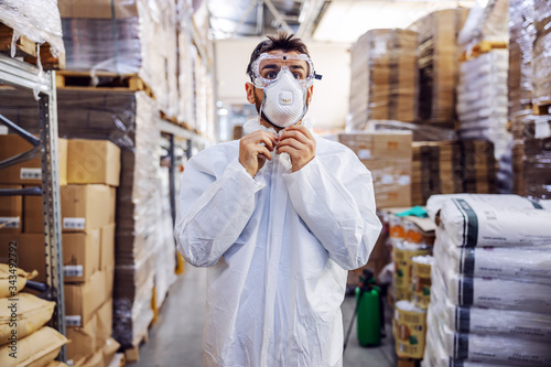 Young man in protective suit putting face mask and preparing himself for disinfection from corona virus / covid-19. Warehouse interior. Warehouse is full of food products.