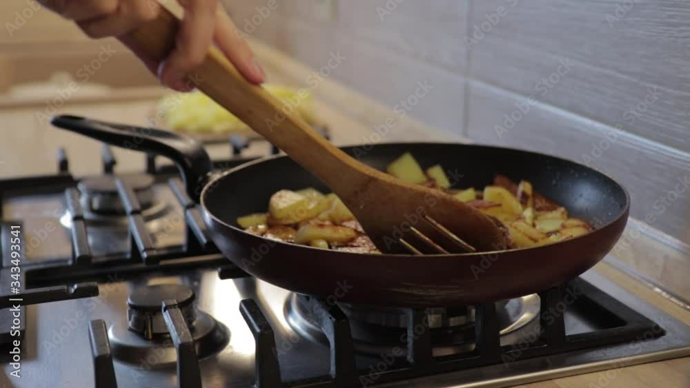 The hand lifts the lid of the pan, stirs the fried potatoes with a wooden spatula on a gas stove