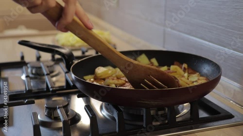 The hand lifts the lid of the pan, stirs the fried potatoes with a wooden spatula on a gas stove