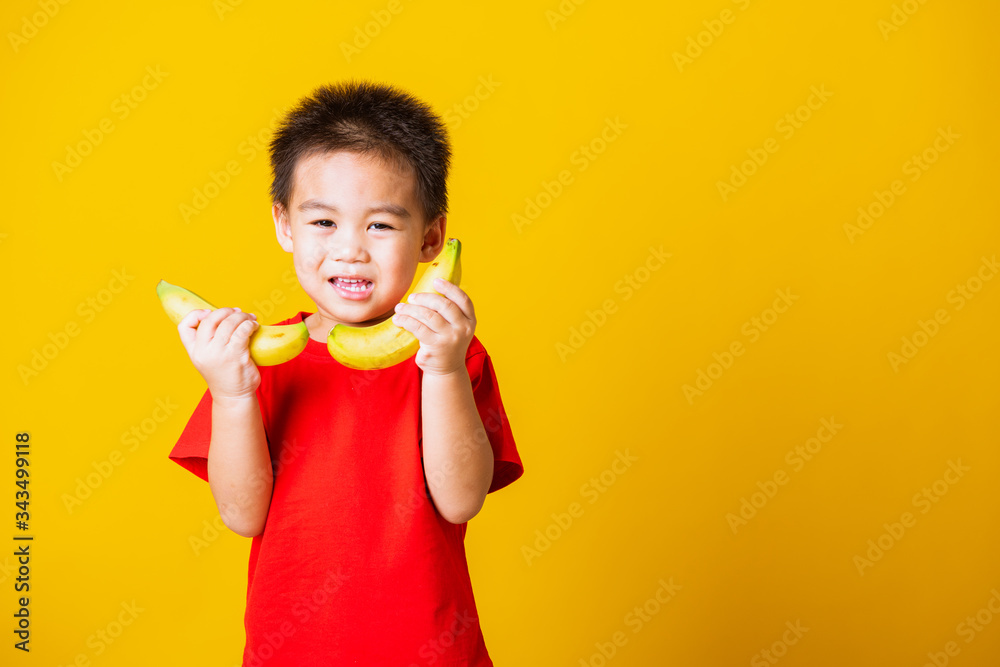 Happy portrait Asian child or kid cute little boy attractive smile wearing red t-shirt playing holds banana fruit, studio shot isolated on yellow background Happy portrait Asian child or kid cute little boy attractive smile wearing red t-shirt playing holds banana fruit, studio shot isolated on yellow background